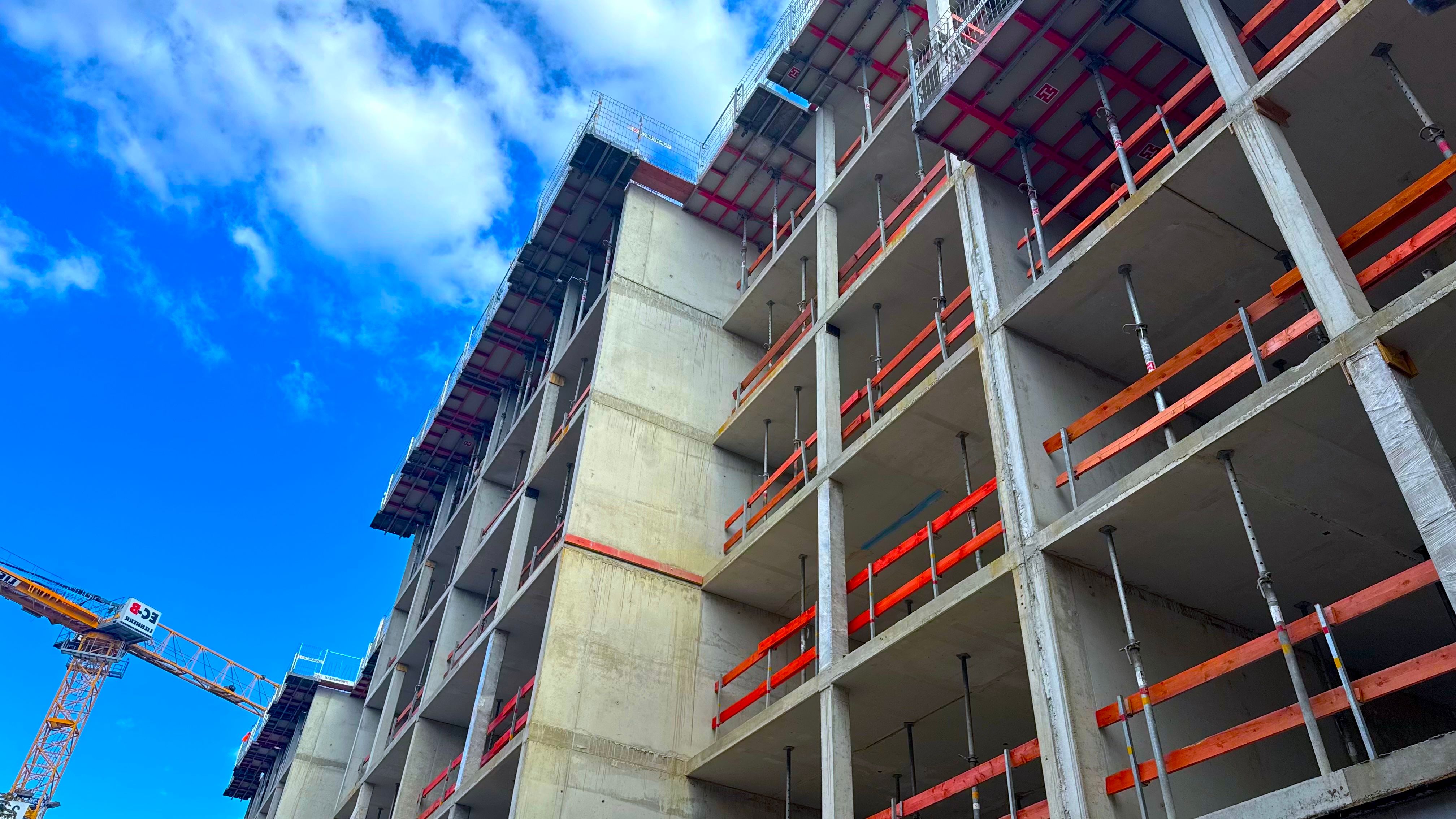 Rohbau eines mehrgeschossigen Wohnhauses aus Stahlbeton mit Deckenstützen und Absturzsicherungen, links ein Baukran vor blauem Himmel.